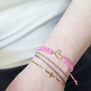 Pink hemp bracelet with gold heart charm worn on wrist with layered gold bracelets.