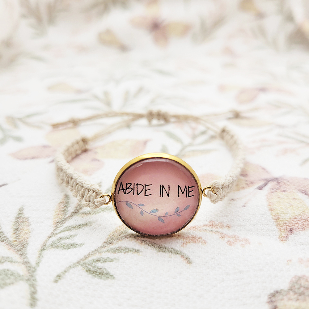 Handmade Abide in Me Christian hemp bracelet with pink glass cabochon charm on floral background.