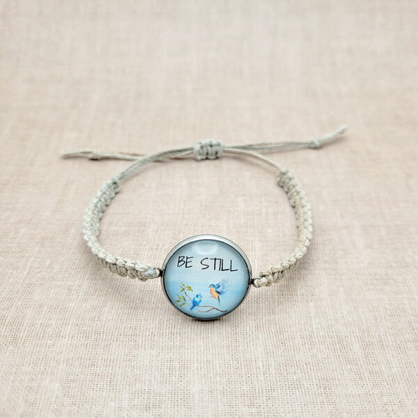 Handmade hemp bracelet with “Be Still” bluebird design on natural linen background, featuring silver bezel and neutral cord