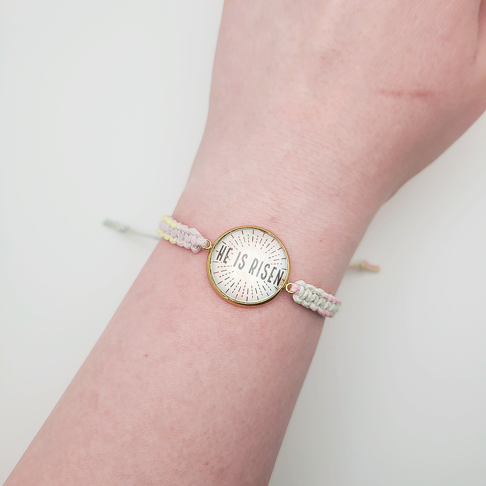 He Is Risen Christian bracelet on pastel hemp cord with gold setting, Easter faith jewelry displayed on wood background