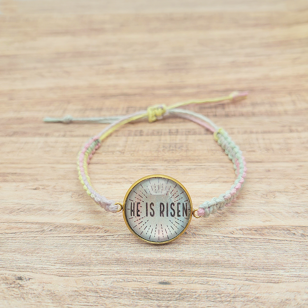 He Is Risen hemp bracelet with pastel multicolor cord and gold bezel on white background, Christian Easter faith jewelry
