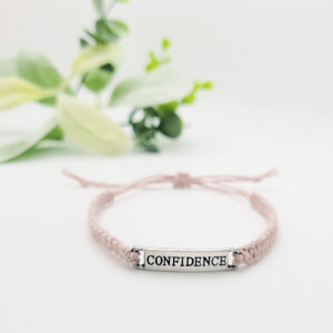 Minimalist adjustable cord bracelet with silver “Confidence” word charm, handmade inspirational bracelet with blush pink cord and soft green plant background.