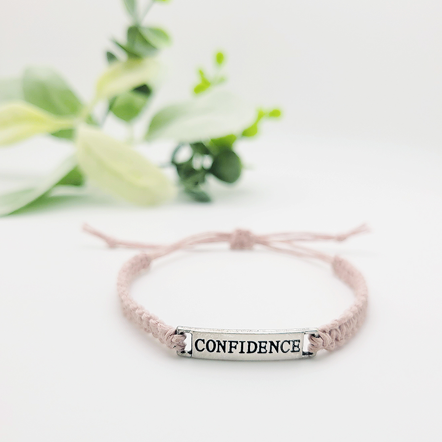 Minimalist adjustable cord bracelet with silver “Confidence” word charm, handmade inspirational bracelet with blush pink cord and soft green plant background.