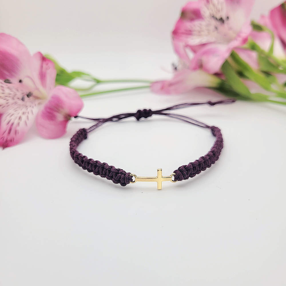 Small gold cross bracelet on burgundy hemp cord with pink flowers in background